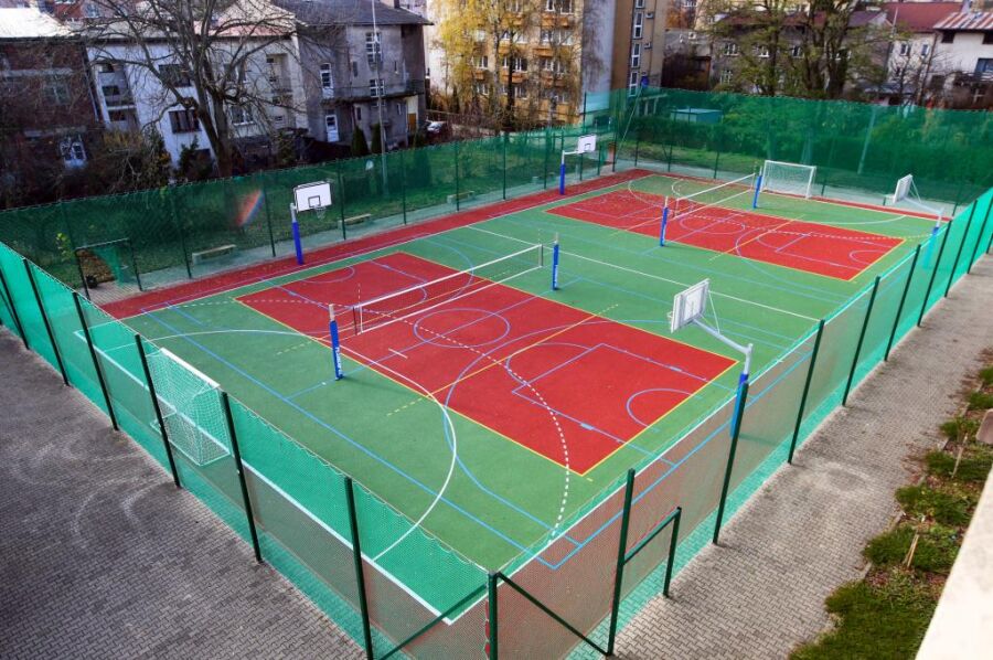 Widok boiska sportowego, z koszami, siatką do siatkówki oraz z wyznaczonymi liniami do gry w tenisa ziemnego. Boisko jest otoczone metalowa siatką.