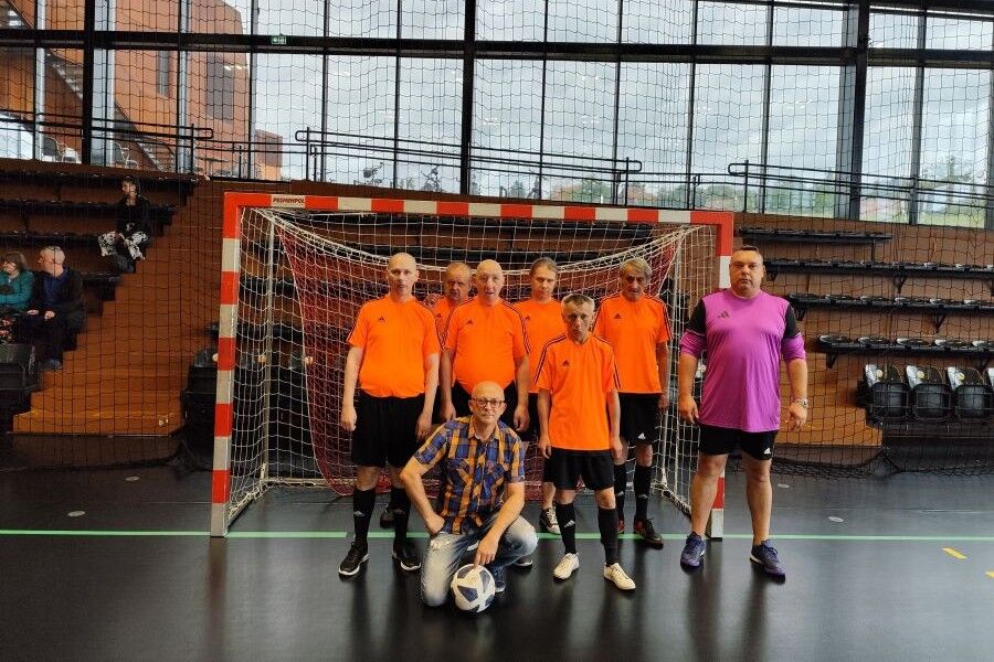 Drużyna w sportowych koszulkach pozuje do wspólnego zdjęcia przed bramką na hali sportowej. Obok zawodników stoi bramkarz i trener.