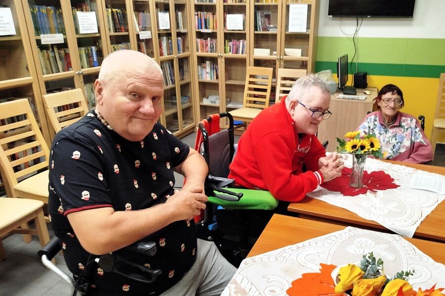 Grupa osób starszych siedzi przy stolikach w pomieszczeniu przypominającym bibliotekę. Na pierwszym planie widać mężczyznę na wózku, obok niego dwie inne osoby również siedzące przy stole.