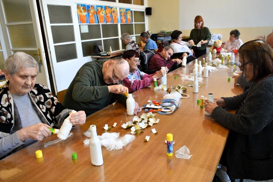 Duża grupa seniorów siedzi wokół długiego stołu i wykonuje prace plastyczne z użyciem butelek, siatki i kleju. Każda osoba skupia się na swojej pracy. Na stole leżą materiały do recyclingu oraz kleje.