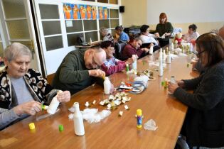 Duża grupa seniorów siedzi wokół długiego stołu i wykonuje prace plastyczne z użyciem butelek, siatki i kleju. Każda osoba skupia się na swojej pracy. Na stole leżą materiały do recyclingu oraz kleje.