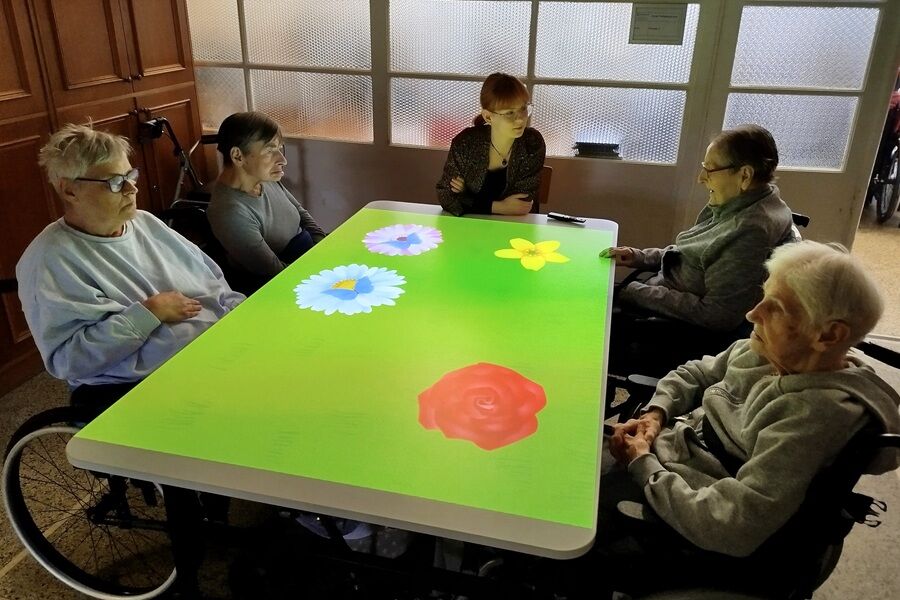 Kilka starszych osób siedzi wokół dużego, podświetlanego stołu interaktywnego, na którego powierzchni wyświetlane są kolorowe kwiaty. Przy stole siedzi także młodsza kobieta, prawdopodobnie prowadząca