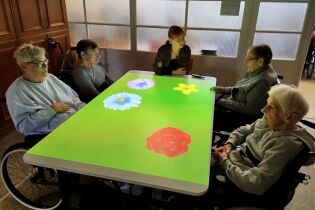 Kilka starszych osób siedzi wokół dużego, podświetlanego stołu interaktywnego, na którego powierzchni wyświetlane są kolorowe kwiaty. Przy stole siedzi także młodsza kobieta, prawdopodobnie prowadząca