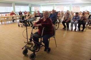 Uśmiechnięta kobieta siedzi na krześle i opiera ręce na chodziku. Za nią znajduje się publiczność siedząca w kilku rzędach w jasnej, przestronnej sali.