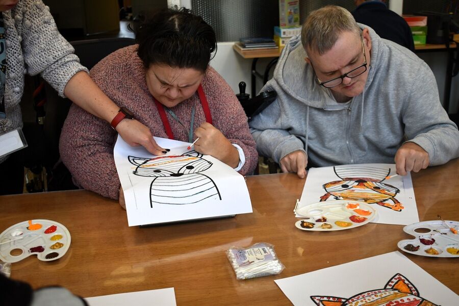 Dwie osoby siedzą przy stole i malują obrazki przedstawiające zwierzę – prawdopodobnie lisa. Po lewej stronie osoba trzyma kartkę i skupia się na malowaniu, a obok niej ktoś pomaga, wskazując miejsce.