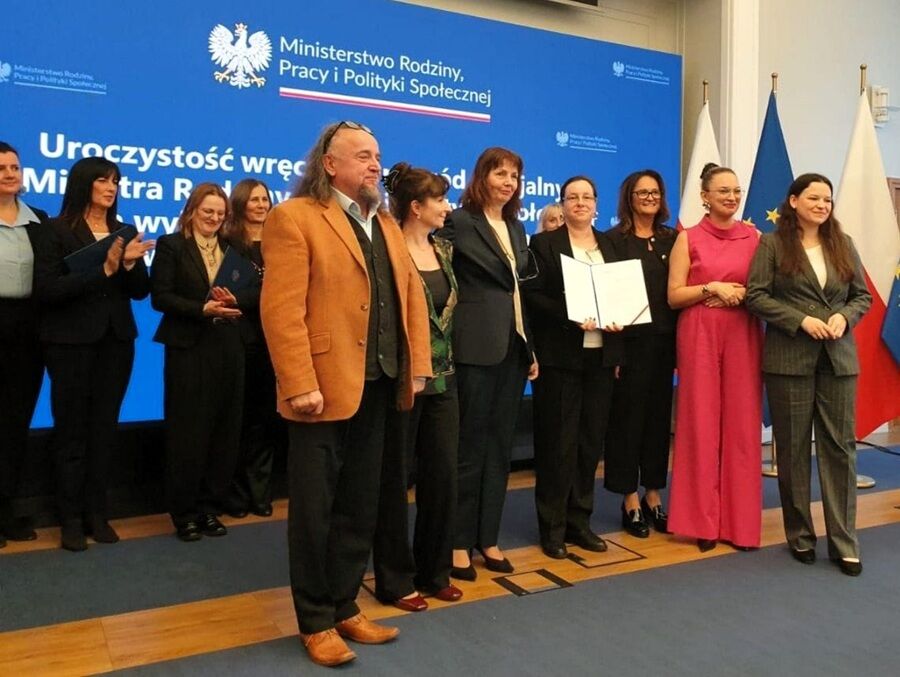 Grupa osób stoi na tle dużej niebieskiej tablicy z logo i napisem „Ministerstwo Rodziny, Pracy i Polityki Społecznej”. W centrum kilka osób pozuje do zdjęcia, a jedna z nich trzyma otwartą teczkę.
