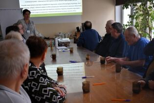 Grupa starszych osób siedzi przy dużym stole i słucha kobiety prowadzącej prezentację wyświetlaną na ekranie. Na stole stoją kubki z herbatą i leżą kolorowe długopisy. Prowadząca stoi przy tablicy.