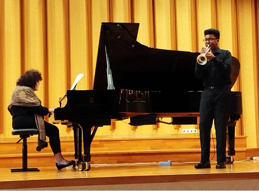 Na scenie w sali koncertowej mężczyzna gra na trąbce, a obok niego kobieta siedzi przy fortepianie i akompaniuje. Oboje są skupieni na występie. W tle znajdują się pionowe, złote panele akustyczne.

