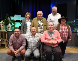 Grupa siedmiu dorosłych osób pozuje do wspólnego zdjęcia na tle sceny z instrumentami i dekoracjami. Jedna kobieta siedzi na wózku, pozostali stoją lub kucają obok niej. Wszyscy uśmiechają się 