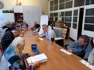 Grupa osób siedzi wokół dużego drewnianego stołu w sali terapeutycznej. W tle widać szafki, regały z materiałami plastycznymi i tablicę.