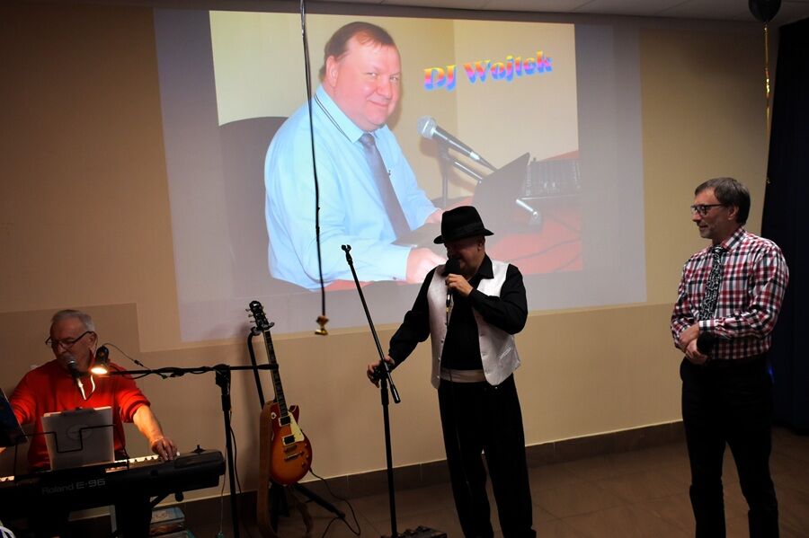 W sali odbywa się występ muzyczny. Na scenie stoi trzech mężczyzn: jeden gra na klawiszach, drugi śpiewa do mikrofonu, a trzeci stoi obok.