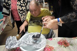 Mężczyzna na wózku inwalidzkim uśmiecha się podczas lania wosku. Trzyma łyżkę z gorącym woskiem razem z drugą osobą. Pod łyżką stoi miska z wodą, a wokół znajdują się osoby uczestniczące w zabawie.

