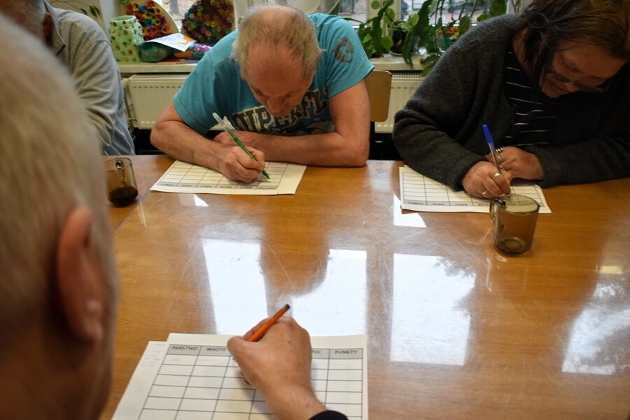 Kilka osób siedzi przy drewnianym stole i pochylając się, zapisuje coś na kartach z tabelami. Na stole stoją kubki z napojami, a w tle widoczne są rośliny na parapecie.

