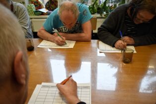 Kilka osób siedzi przy drewnianym stole i pochylając się, zapisuje coś na kartach z tabelami. Na stole stoją kubki z napojami, a w tle widoczne są rośliny na parapecie.

