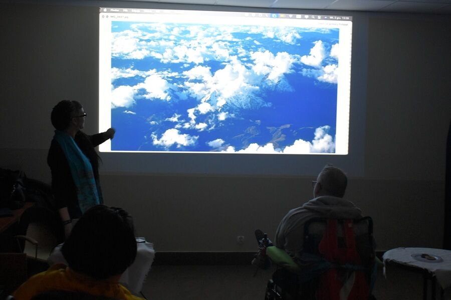 Kobieta stoi przy ekranie projekcyjnym w zaciemnionym pomieszczeniu i wskazuje ręką fotografię chmur oraz oceanu. Kilka osób siedzi przed nią i ogląda prezentację.