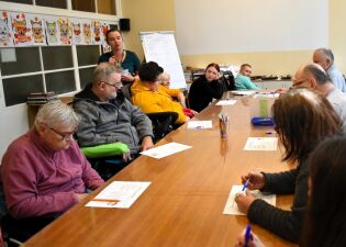 Grupa dorosłych osób siedzi przy długim stole w sali warsztatowej. Uczestnicy pracują z kartkami i długopisami, a prowadząca stoi na końcu stołu i tłumaczy zadanie. 