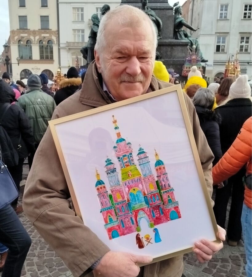 Starszy mężczyzna stoi na tle tłumu na miejskim rynku i trzyma w rękach oprawiony obraz przedstawiający kolorową, wielowieżową szopkę krakowską. Ubrany jest w brązową kurtkę. 