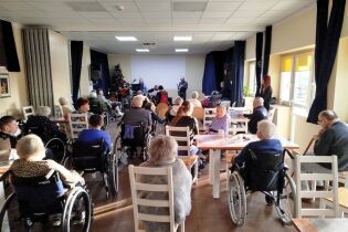 W jasnej sali zgromadzeni są seniorzy oraz osoby z niepełnosprawnościami, siedzący na krzesłach i w wózkach inwalidzkich. Na scenie kilka osób gra na instrumentach i śpiewa. 
