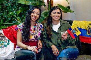 Priscilla Bravo i Valeria Olivas. Zdjęcie dwóch młodych kobiet siedzących w pomieszczeniu biurowym na sofie. W tle zielone rośliny doniczkowe. Obok kobiet leżą flagi narodowe Ekwadoru i Meksyku. 