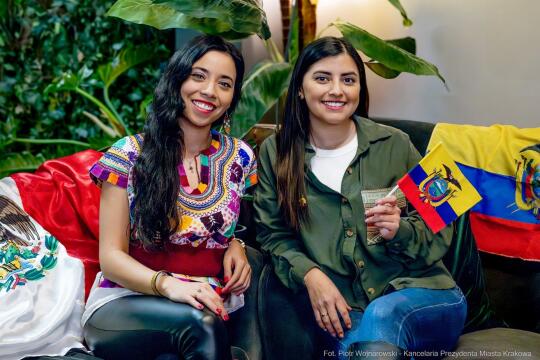 Priscilla Bravo i Valeria Olivas. Zdjęcie dwóch młodych kobiet siedzących w pomieszczeniu biurowym na sofie. W tle zielone rośliny doniczkowe. Obok kobiet leżą flagi narodowe Ekwadoru i Meksyku. 