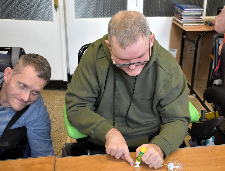 Dwóch mężczyzn przy stole podczas zajęć plastycznych – jeden nakłada klej na mały element, w tle pomieszczenie świetlicy.