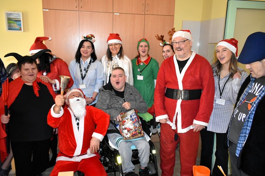 Grupa osób w strojach świątecznych i czapkach Mikołaja pozuje do wspólnego zdjęcia w pomieszczeniu, atmosfera radosna i świąteczna.