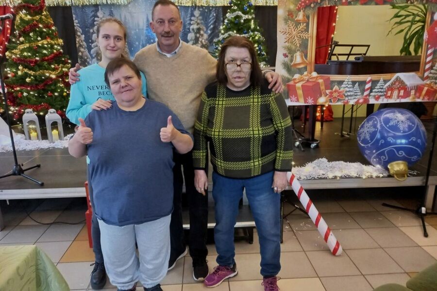 Cztery osoby stoją przed świąteczną dekoracją z choinkami i bombkami, dwie osoby pokazują uniesione kciuki do góry.