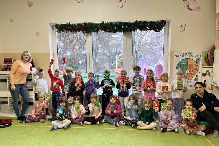 grupa dzieci siedzących i stojących na zielonym dywanie w sali przedszkolnej.