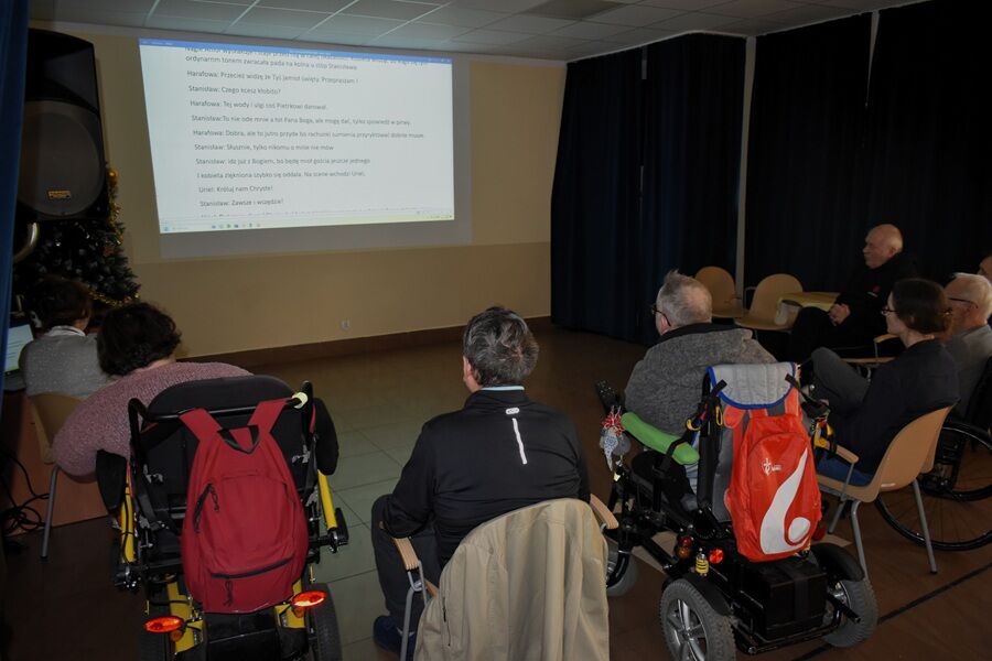 Grupa osób, w tym kilka osób na wózkach inwalidzkich, siedzi w sali i ogląda tekst wyświetlany na dużym ekranie. W pomieszczeniu panuje półmrok, a uczestnicy są zwróceni w stronę ekranu.