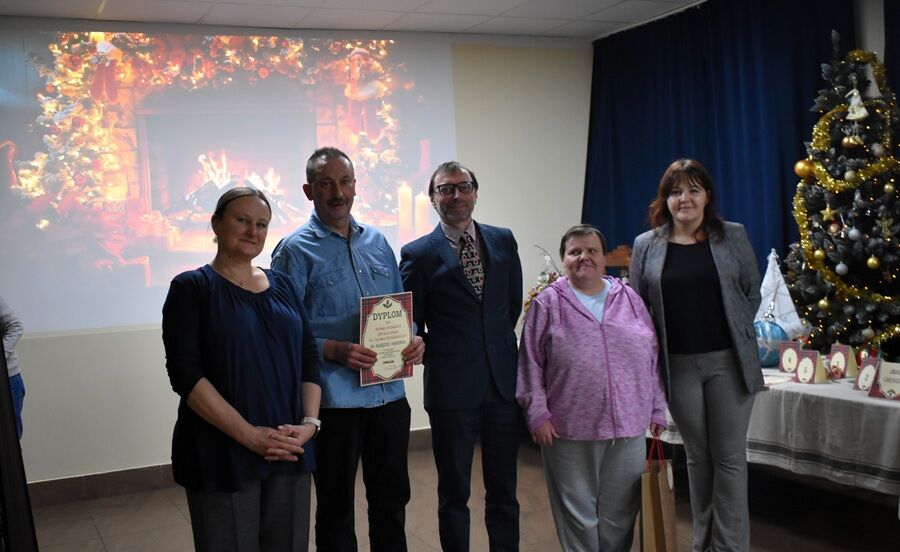 Pięć osób stoi razem w sali przed świąteczną dekoracją z kominkiem wyświetlonym na ekranie. Jedna osoba trzyma dyplom. Obok stoi ozdobiona choinka i stół z nagrodami.

