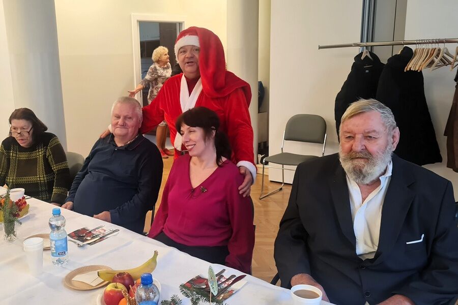 Kilka osób siedzi przy świątecznie nakrytym stole. Za jedną z par stoi osoba w stroju Mikołaja, która obejmuje ich ramionami. Na stole znajdują się owoce, napoje i dekoracje świąteczne.

