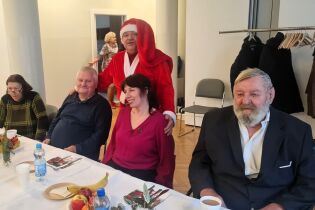 Kilka osób siedzi przy świątecznie nakrytym stole. Za jedną z par stoi osoba w stroju Mikołaja, która obejmuje ich ramionami. Na stole znajdują się owoce, napoje i dekoracje świąteczne.

