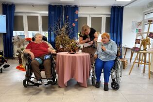 Na zdjęciu Pani prowadząca zadaje pytania odnośnie pracy konkursowej, mieszkanki siedzą na wózkach przy stoliku, na którym wyeksponowana jest praca konkursowa.