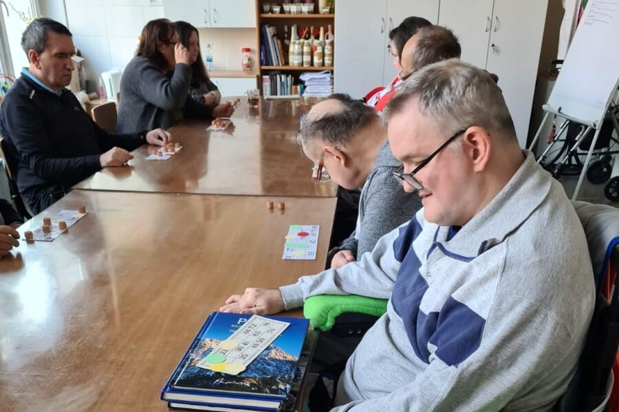 Grupa dorosłych osób siedzi przy długim stole w świetlicy, grając w grę planszową z kartami i beczułkami; uczestnicy skupieni są na rozgrywce, a na stole leżą plansze i beczułki.