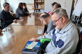 Grupa dorosłych osób siedzi przy długim stole w świetlicy, grając w grę planszową z kartami i beczułkami; uczestnicy skupieni są na rozgrywce, a na stole leżą plansze i beczułki.