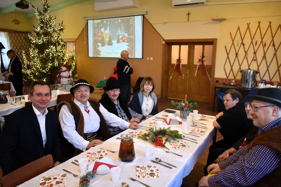 Uczestnicy spotkania świątecznego siedzą przy udekorowanym stole z opłatkiem; w tle stoi choinka i świąteczne dekoracje, a atmosfera jest uroczysta i wspólnotowa.