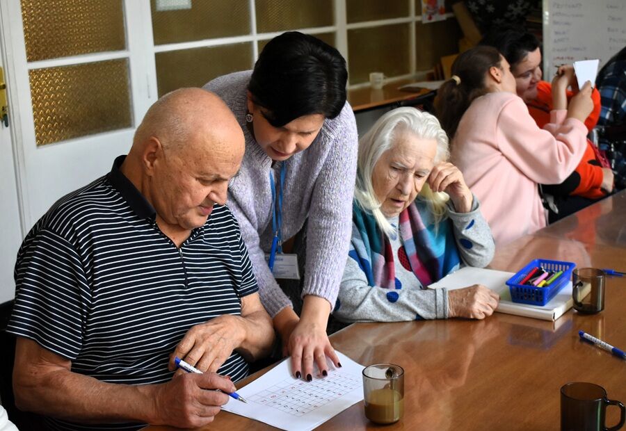 Starsze osoby pracują przy stole nad zadaniem papierowym; opiekunka pomaga jednej z nich, wskazując na kartę, a na stole znajdują się długopisy i kubki z napojami.