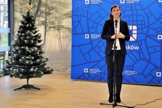 kobieta stojąca obok udekorowanej świątecznie choinki przemawia przy użyciu mikrofonu na statywie