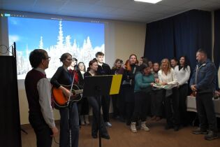 Grupa dorosłych osób stoi w sali i wspólnie śpiewa; jedna osoba gra na gitarze przy mikrofonie. W tle wyświetlany jest zimowy krajobraz na ekranie.