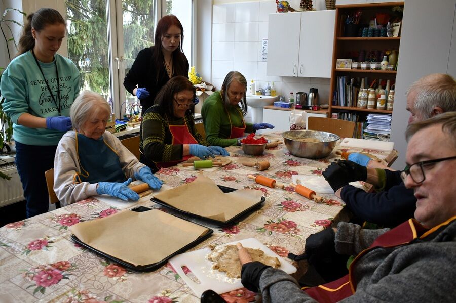 Kilka starszych osób siedzi przy stole i wspólnie przygotowuje wypieki, wałkując ciasto i układając je na blachach; obok stoją opiekunowie pomagający w pracy.