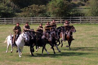 Grafika przedstawia grupę osób na koniach, w trakcie rekonstrukcji bitwy.