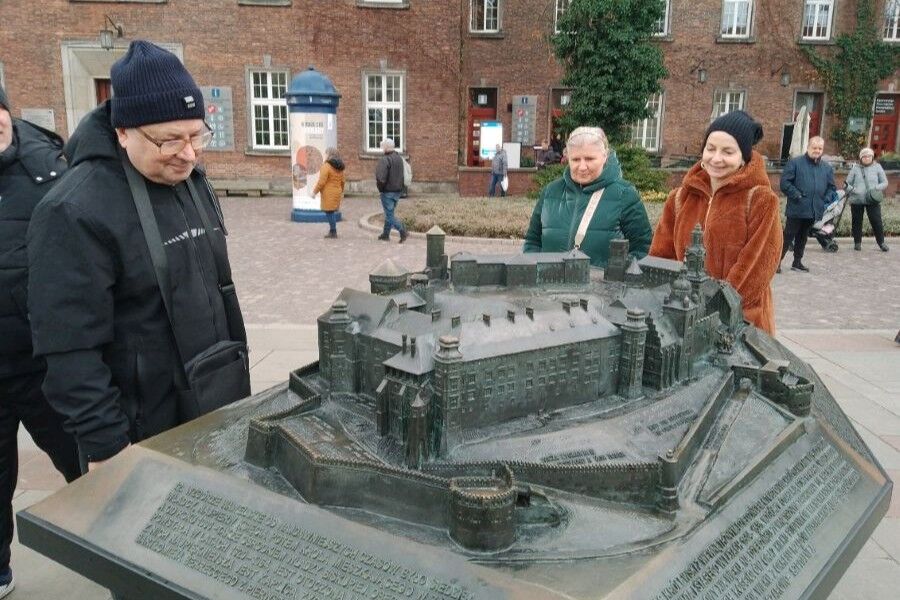 Dwie kobiety i mężczyzna patrzą na model wzgórza wawelskiego. Tło obrazu stanowią historyczne budynki i przestrzeń miasta.