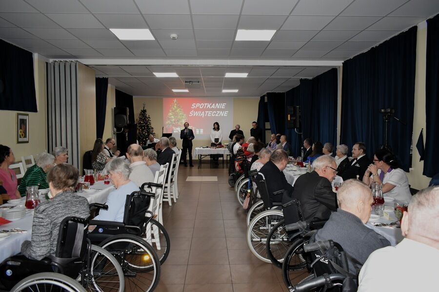 Sala z długimi stołami, przy których siedzą uczestnicy spotkania świątecznego, w tym osoby poruszające się na wózkach inwalidzkich; z przodu sali stoją prowadzący, a na ekranie wyświetlony jest napis 