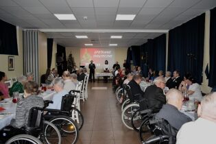 Sala z długimi stołami, przy których siedzą uczestnicy spotkania świątecznego, w tym osoby poruszające się na wózkach inwalidzkich; z przodu sali stoją prowadzący, a na ekranie wyświetlony jest napis 