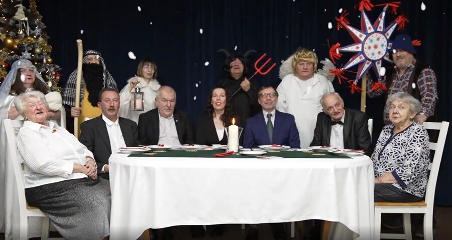 Grupa osób siedzi przy wspólnym stole przykrytym białym obrusem; uczestnicy są przebrani w stroje nawiązujące do jasełek, na stole pali się świeca, a w tle widać choinkę oraz świąteczne dekoracje.