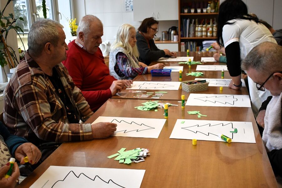 Kilka osób siedzi przy stole i wykonuje prace plastyczne z papieru, wycinając kształty choinek; na stole kleje, papier i materiały do rękodzieła.