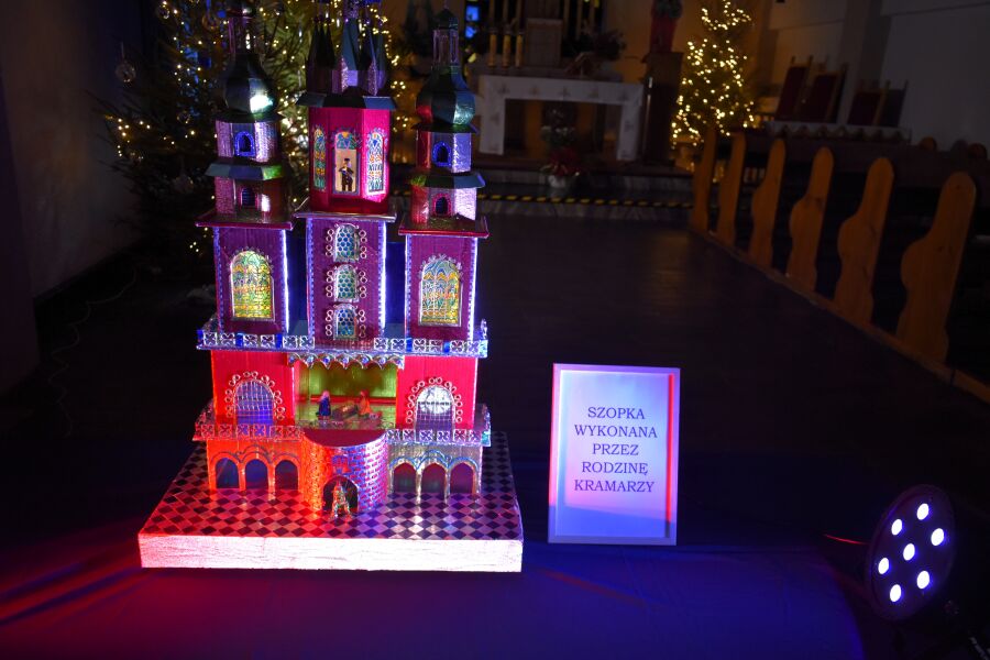 Kolorowa, podświetlana szopka krakowska z wieżyczkami i witrażowymi oknami, ustawiona w kościele; obok tabliczka z informacją o autorach, w tle świąteczne dekoracje.