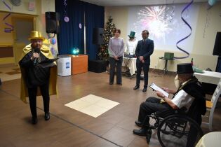 Grupa osób podczas sylwestrowego wydarzenia w sali: jedna osoba przemawia do mikrofonu, inni stoją i siedzą, w tle choinka, dekoracje oraz projekcja fajerwerków.