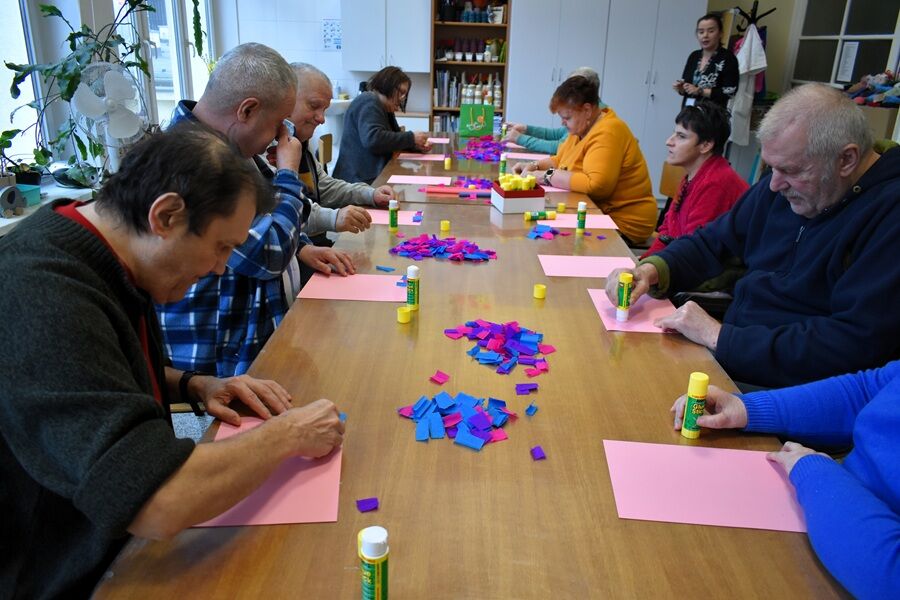 Grupa dorosłych osób siedzi przy długim stole i wykonuje prace plastyczne z kolorowego papieru i kleju. Uczestnicy są skupieni na rękodziele, a na stole leżą rozłożone materiały artystyczne.