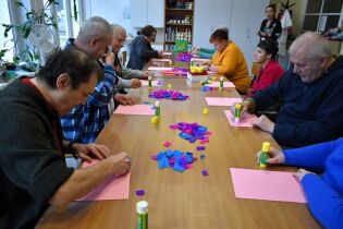 Grupa dorosłych osób siedzi przy długim stole i wykonuje prace plastyczne z kolorowego papieru i kleju. Uczestnicy są skupieni na rękodziele, a na stole leżą rozłożone materiały artystyczne.